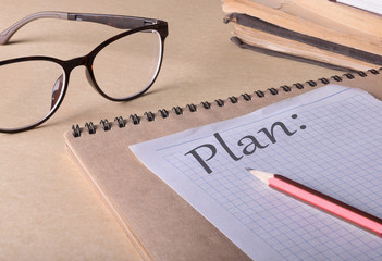 Paper note written with PLAN  inscription.  Eye glasses, book and pencil on table.- image
