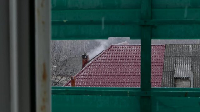 Smoking chimneys at roof with snow of hous. It is snowing. Winter day, heating season. Shot over a green fence. 4K 2160p 25fps UHD video.