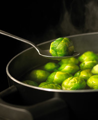 SPOON HOLDING BRUSSELS SPROUT OVER PAN OF BOILING WATER FULL OF SPROUTS WITH RISING STEAM SIMMERING ON GAS COOKER