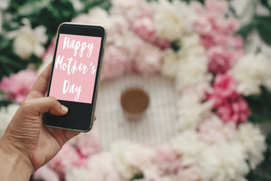 Happy Mother's Day text sign on screen phone in hand at  stylish pink and white peonies on pastel pink paper, flat lay. Stylish floral greeting card.