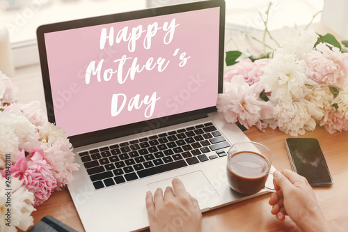 Happy Mother's Day text sign on pink laptop screen and girl hands with coffee, phone, black notebook and peonies on rustic wooden table. Stylish floral greeting card.