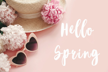 Hello Spring text sign on stylish girly pink retro sunglasses, white and pink peonies, straw hat on pastel pink paper, flat lay. Stylish floral greeting card.