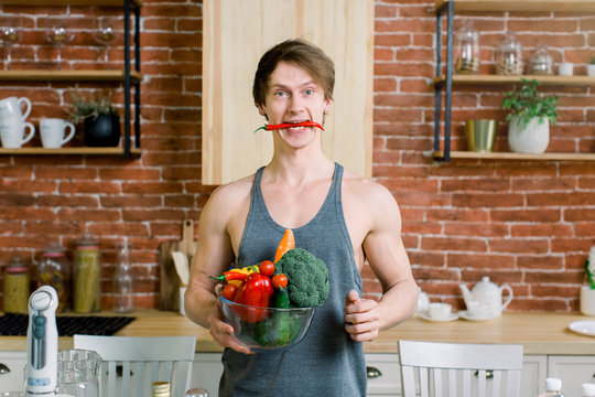 Portrait Of A Handsome Sports Man Holding Glass Bowl With Fresh Vegetables And Fruits, Red Hot Chili Pepper In His Mouth On The Kitchen At Home