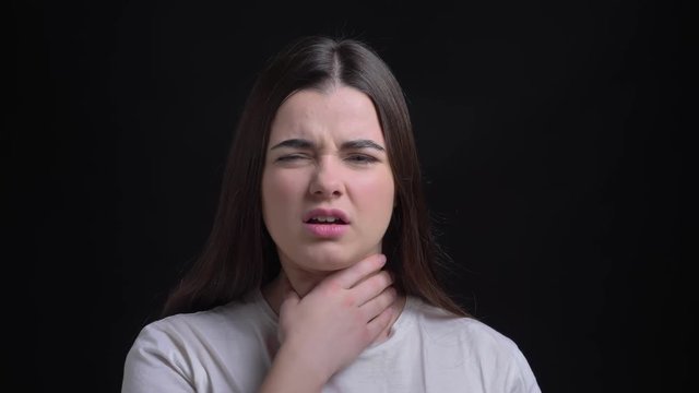 Portrait of young overweight brunette caucasian girl having sore throat and touching her neck on black background.