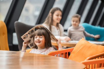 A charming little girl was sitting at a wooden table with a menu on her head in the restaurant