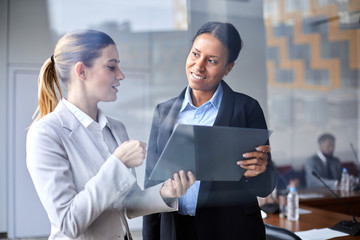 Two young intercultural businesswomen discussing online data for report in conference hall