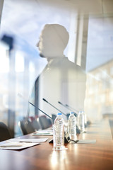 Row of plastic bottles of water, chairs, microphones and papers on table and reflection of speaker...
