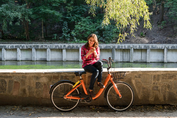 Subject ecological bicycle transport. Young Caucasian woman in shirt student sits resting in a park near the lake for rent orange bike uses a mobile phone autumn break in sunny weather technology