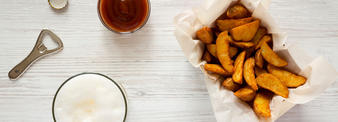 Fried potato wedges in paper box, barbecue sauce and beer on a white wooden table, top view. Overhead, flat lay, from above.