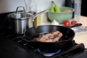 New York strip steak frying in a cast iron pan on a natural gas stove top.