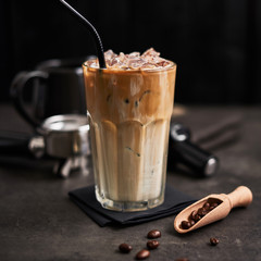 Ice coffee with cream in a tall glass and coffee beans, portafilter, tamper and milk jug on dark...