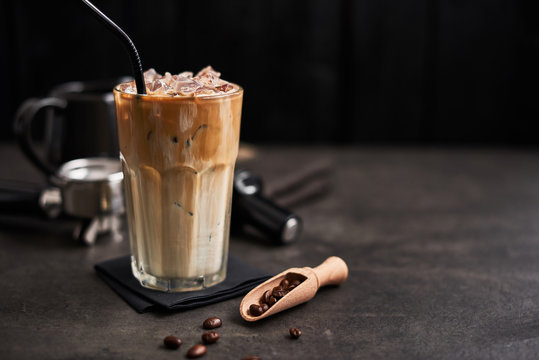 Ice coffee with cream in a tall glass and coffee beans, portafilter, tamper and milk jug on dark concrete table over black wooden background. Cold summer drink. Copy space for text. Selective focus.