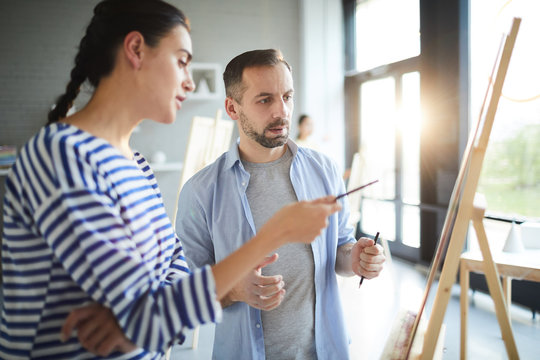 Young woman consulting with teacher of painting about her idea while pointing at easel