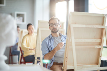 Fototapeta premium Serious student of faculty of arts painting human head while sitting in front of easel at lesson