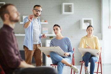 Two concentrated learners looking at their groupmate while going to sketch him at lesson of painting