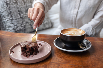 A woman cutting brownie cake with fork with coffee cup on wooden table in cafe