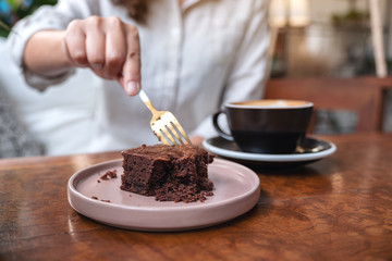 A woman cutting brownie cake with fork with coffee cup on wooden table in cafe