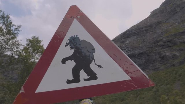 Red and white triangle street sign funny with a troll under a bridge in a mountainous landscape with cliff and trees on an overcast day