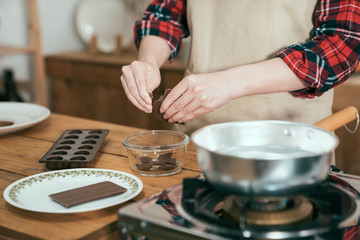 close up woman hands photo peels chocolate into small pieces putting in bowl with boiled water in the pot on stove in kitchen. female in apron handmade dessert on valentines day for lover boyfriend.