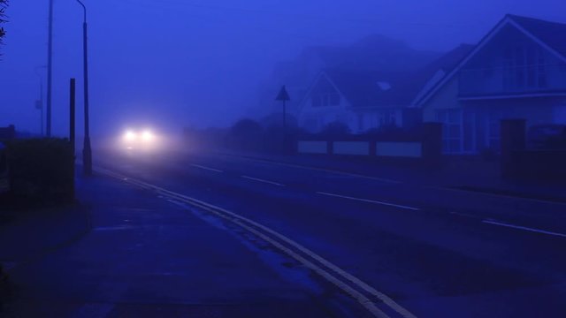 Foggy morning in UK by the coast. Dark blue hue. Street lights are still on. Car, with headlights on, is driving towards the camera empty road and disappears off sight. Nobody else is visible. HD