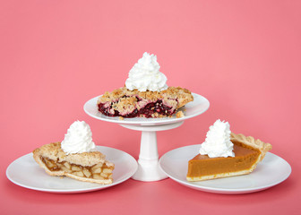 Apple and pumpkin pie on plates with whipped cream, cherry pie on plate with whipped cream on pedestal above them. Pink background. National pie day march 14