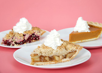 Close up on apple, cherry and pumpkin pie slices on individual plates with whipped cream on top of them on pink background. Focus on front pie, apple. National pie day march 14