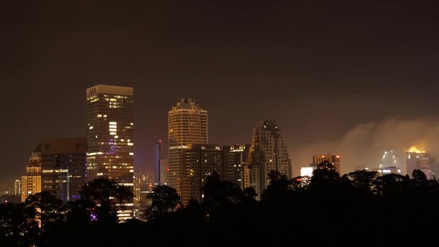 Night Time Timelapse Over View Of Kuala Lumpur Skyline Illuminated Buildings & Flight Path Of Plane Against Fast Clouds.