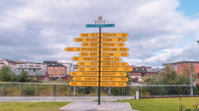 Timelapse zooming in and out from a large yellow sign pointing the direction of many famous cities around the world on a sunny day in a park by the city