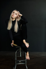 Beautiful blonde in a black suit on a black background sitting on a chair.Makeup, cosmetics, hair care. Fashionable clothes. Fashion, Beauty, style.