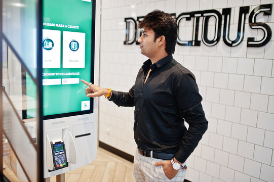 Indian man customer at store place orders and pay through self pay floor kiosk for fast food, payment terminal. Make a choise of language on screen.