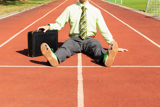 An exhausted businessman in gray suit with green shirt and tie, black briefcase and broken green running shoes sitting on a running track