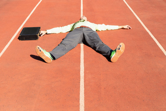 An exhausted businessman in gray suit with green shirt and tie, black briefcase and broken green running shoes lying on a running track