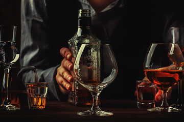 The bartender holding bottle of strong drink in hand and big wine glass on the old bar counter. Vintage wooden background in pub or bar, night mood. Place for text, toning, selective focus