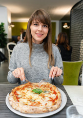 Woman eats with knife and fork a pizza Margherita with mozzarella tomatoes and basil. Neapolitan pizza from wood-burning stove. lunch in an Italian restaurant. Table near to a large window.
