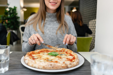 Woman eats with knife and fork a pizza Margherita with mozzarella tomatoes and basil. Neapolitan pizza from wood-burning stove. lunch in an Italian restaurant. Table near to a large window.