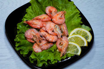 Boiled shrimps with lemon slices on a sheet of lettuce on a black plate on a wooden background.
