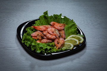 Boiled shrimps with lemon slices on a sheet of lettuce on a black plate on a wooden background.
