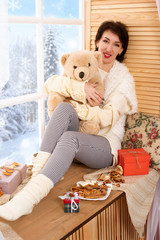 Woman is sitting with bear toy on a window sill. Beautiful view outside the window - sunny day in winter forest and snow.