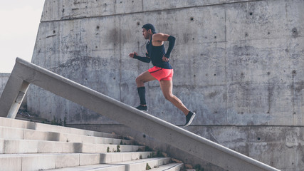 Fototapeta premium Muscular hispanic Young male dark-skinned athlete running up a flight of stairs with speed, sporty young man in black t-shirt training or working out outdoors while jogging up the steps.
