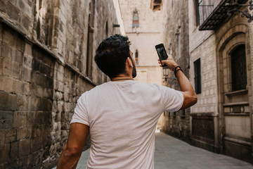 Obraz na płótnie Canvas Content attractive hispanic man in white tshirt photographing himself on mobile phone while enjoying city travel.Bearded hipster male making selfie via smartphone on gotic streets