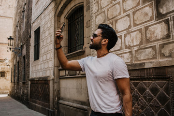 Fototapeta na wymiar Content attractive hispanic man in white tshirt photographing himself on mobile phone while enjoying city travel.Bearded hipster male making selfie via smartphone on gotic streets