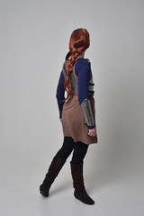 full length portrait of a  red haired girl wearing medieval warrior costume and steel armour, standing pose with back to the camera on grey studio background.