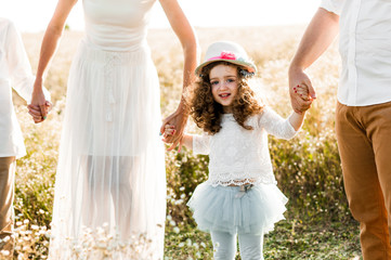 Happy curly girl in a hat holding parents hands in the midst of a chamomile field