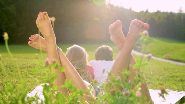 SLOW MOTION, LENS FLARE, CLOSE UP, DOF: Unrecognizable barefoot couple lying on the blanket and watching the picturesque nature. Summer sun rays shining on newlyweds lying in the meadow during a date.