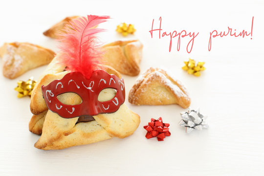 Purim celebration concept (jewish carnival holiday). Traditional hamantaschen cookies with cute mask over white wooden table.