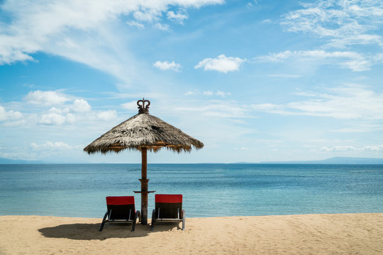 Two Red Beach Chairs Under Wicker Unbrella On A Coast.