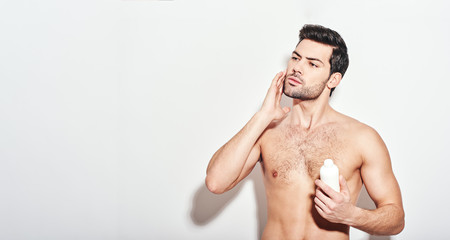 Another beauty treatment. Handsome man applies the moisturizer standing isolated over white...