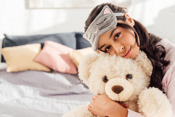 beautiful african american girl in sleeping mask hugging teddy bear and looking at camera in bedroom