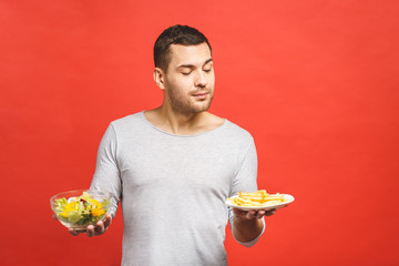 Portrait of young handsome man craving after junk food instead of eating healthy food, isolated over red background.