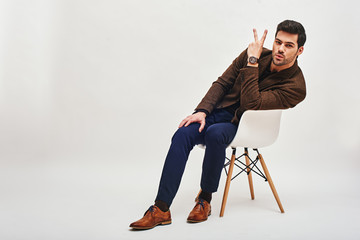 Victory! Stylish dark-haired young man sitting on a chair, leaning back and showing 'V' sign isolated over white background